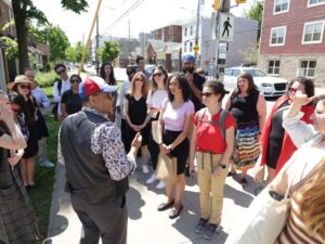 Un homme se tient sur le trottoir et s'adresse à un grand groupe de personnes qui l'écoutent attentivement.